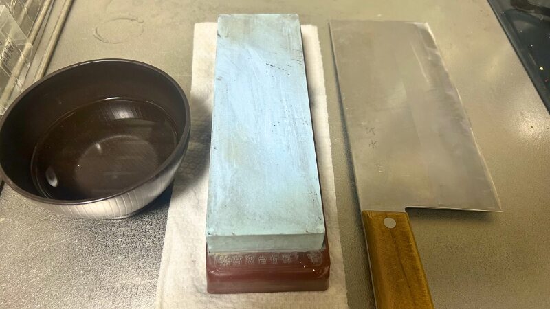 お椀に入れた水を指先ですくい、砥石の表面を濡らしながら中華包丁を研ぐ準備をする様子。研ぎ汁(泥)を保つためのプロの工夫