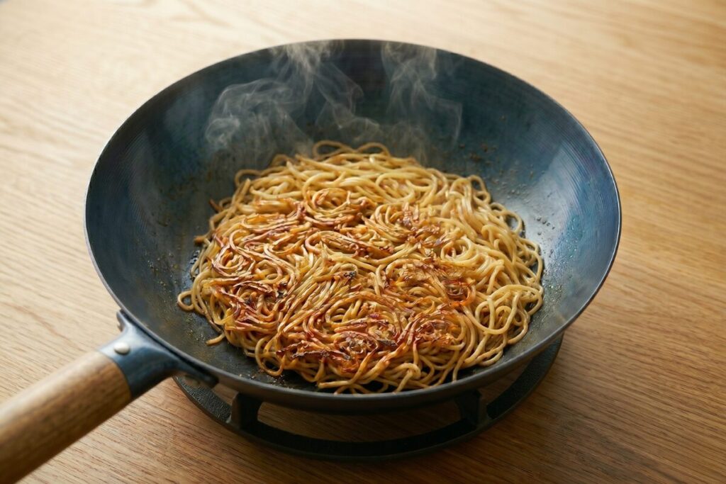 中華鍋で焼きそばの麺を「揚げ焼き」にしている様子。底面にこんがりと香ばしい焼き色がつき、湯気が上がっている。