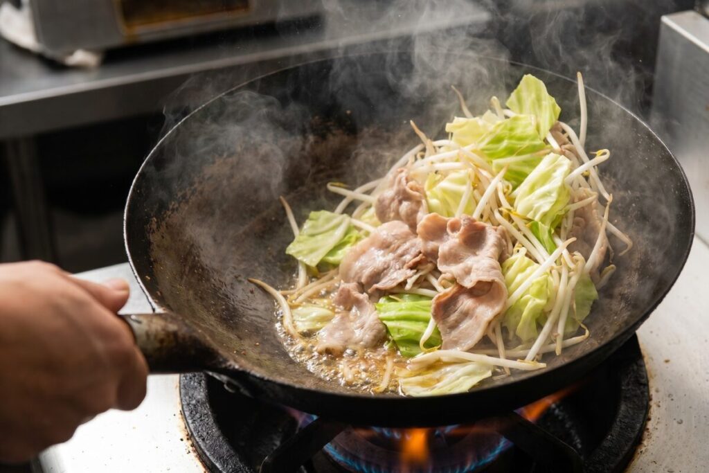 中華鍋で豚肉とキャベツ、もやしを強火で一気に炒めている躍動感のある写真。湯気が立ち上り、野菜のシャキシャキ感が伝わる仕上がり。