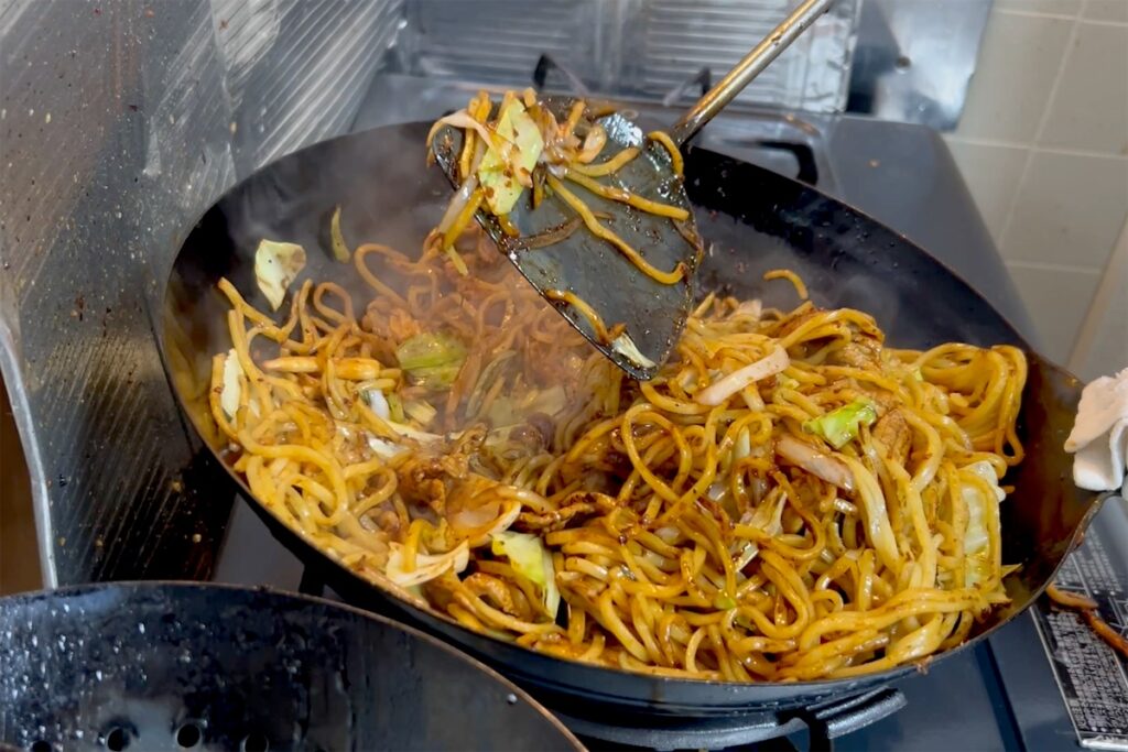 中華ヘラで焼きそばの麺とキャベツを持ち上げ、大きく返すところ
