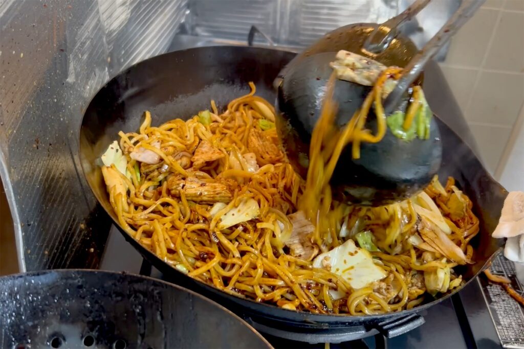 ヘラとお玉で焼きそばの麺を挟み、持ち上げて混ぜるところ
