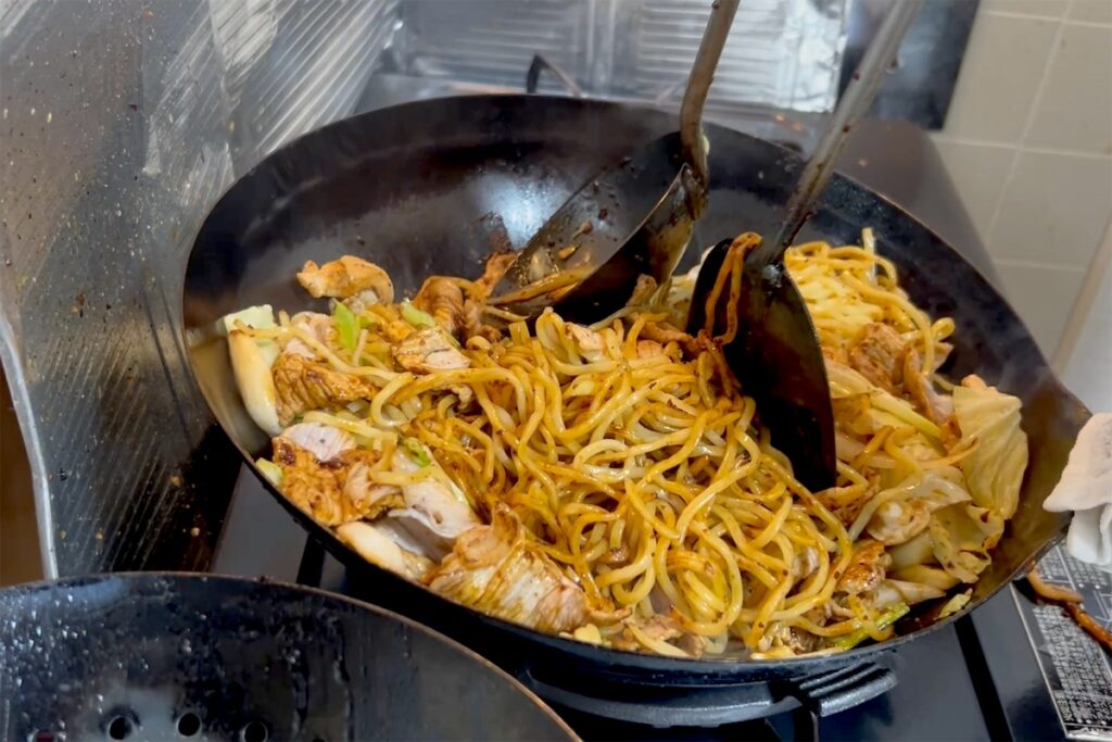 コンロに置いた中華鍋を振らずに、ヘラとお玉の2本で焼きそばを混ぜるところ